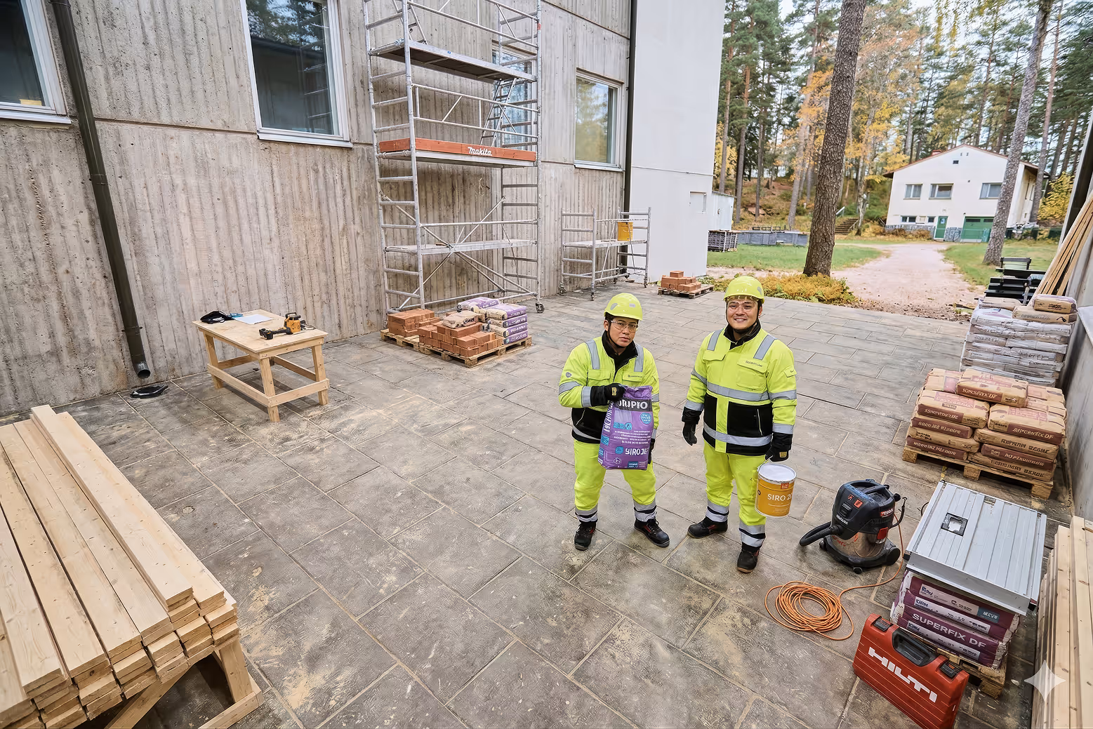 Kaksi työmiestä keltaisissa suojavaatteissa ja kypärissä rakennustyömaalla ulkona.