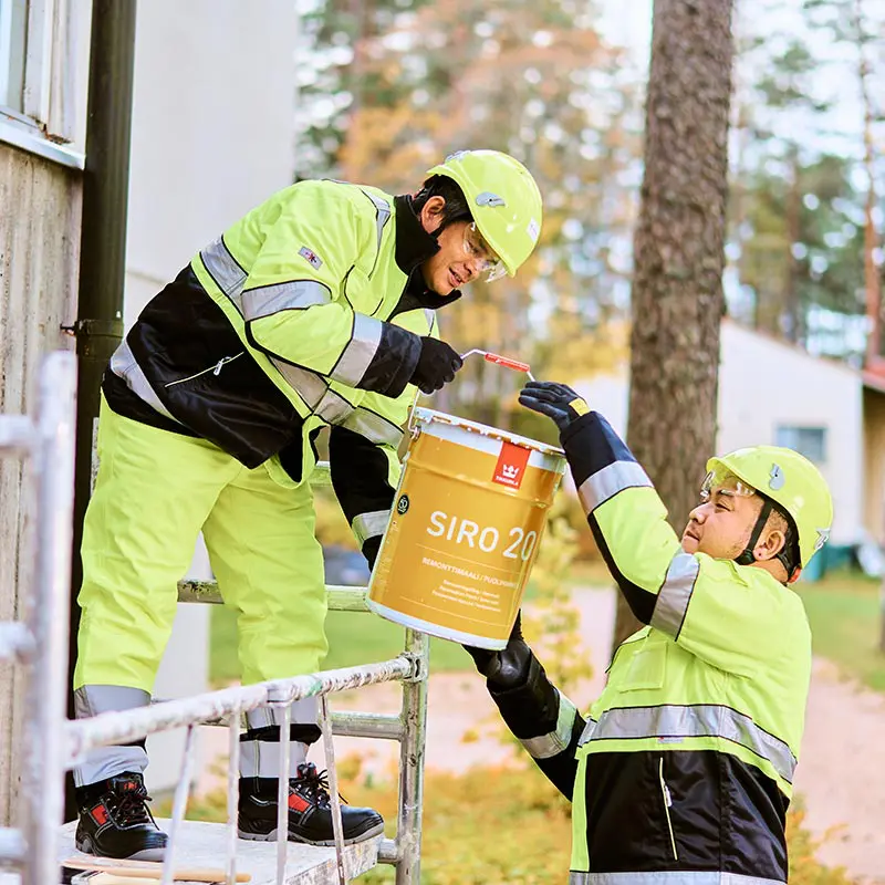 Kaksi rakennustyöntekijää keltaisissa suojavaatteissa vaihtaa maalaustarvikkeita ulkona.