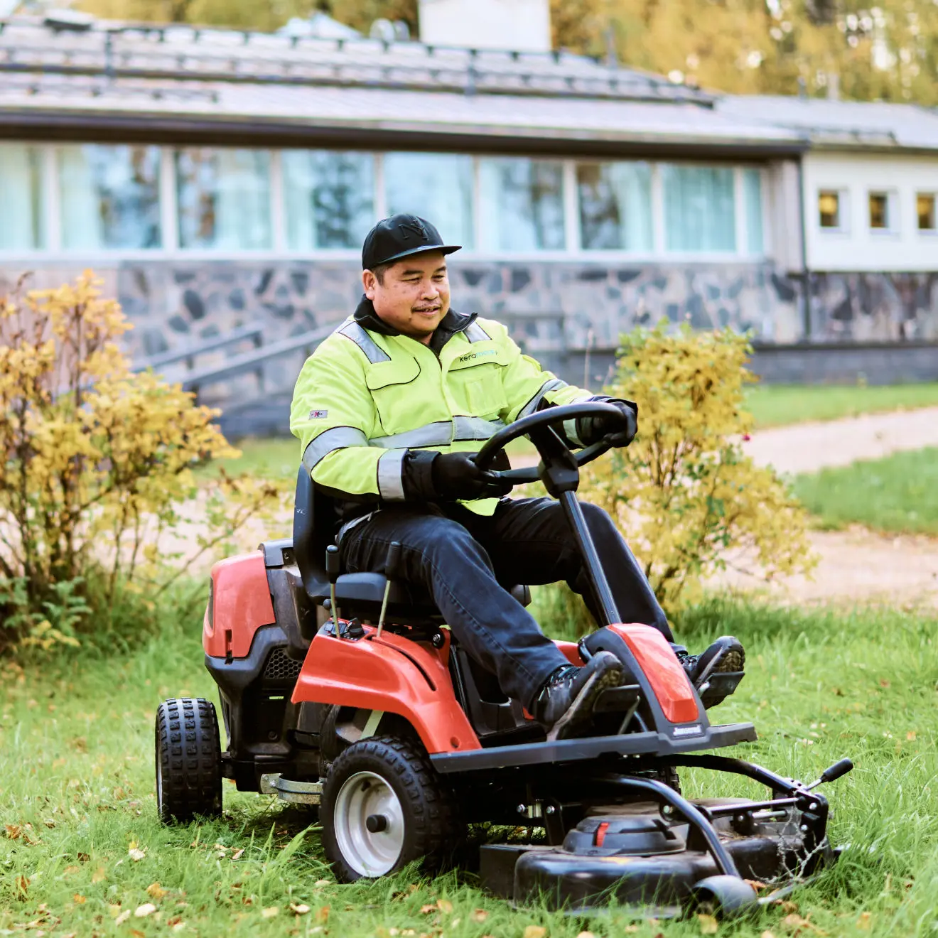 Mies ajaa vihreällä nurmikolla oranssia ruohonleikkuria pihatöissä talon edustalla.