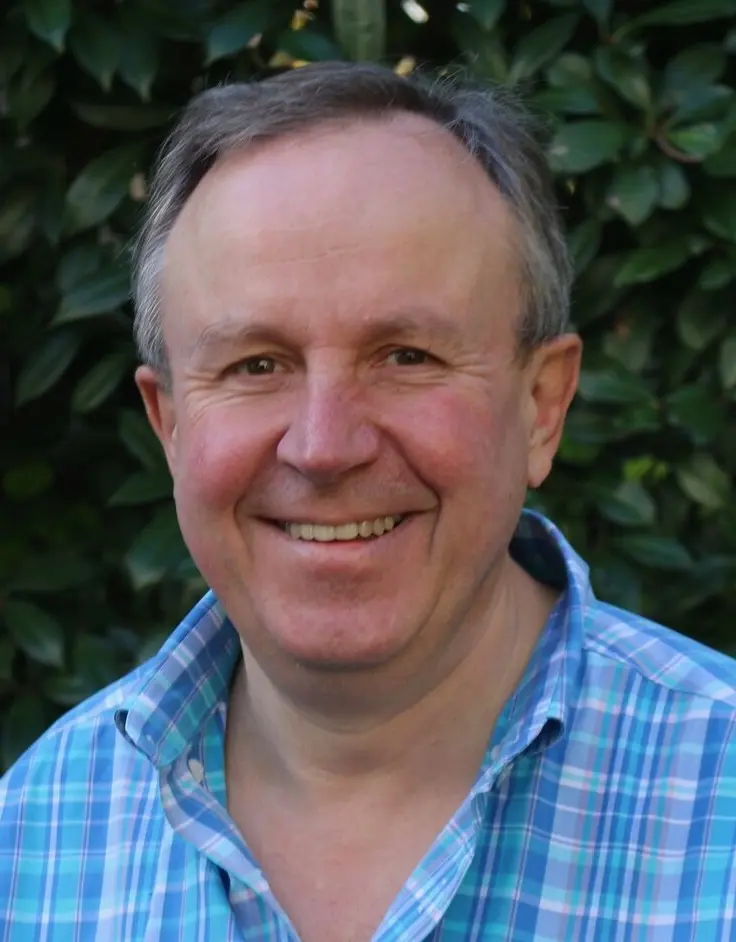 Middle-aged man smiling outdoors in a blue checkered shirt with greenery background.