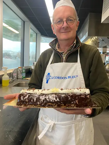 Man in Le Cordon Bleu apron holding a chocolate yule log cake in a kitchen.