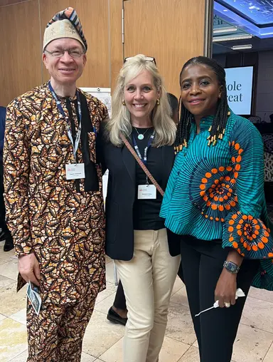 Three people smiling indoors, two wearing colorful African prints and one in a black blazer and beige pants.