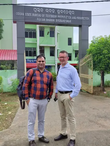 Two men standing on a path in front of Odisha State Bureau of Textbook Preparation and Production building.
