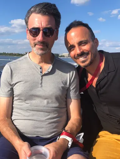 Two men sitting outdoors by the water under a blue sky with clouds, smiling and wearing casual clothes.