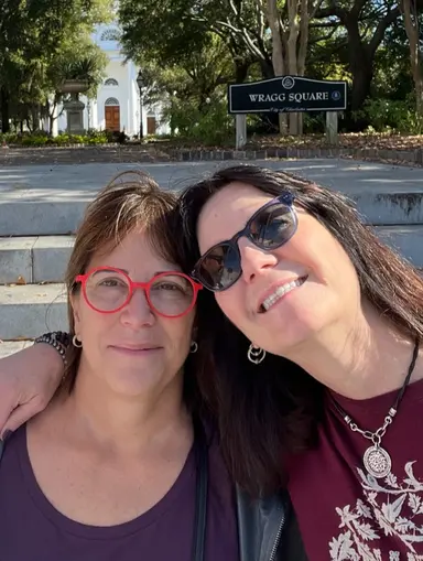 Two women smiling, one with red glasses and one with sunglasses, sitting outdoors near Wragg Square sign.