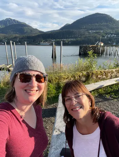 Two smiling women outdoors near a lake with dock and forested mountains in the background.