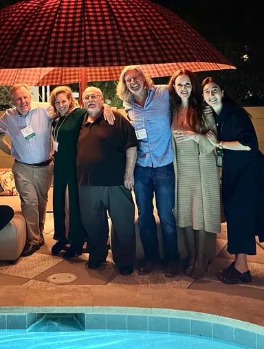 Six diverse adults smiling and standing close together by a pool under a large checkered umbrella at night.