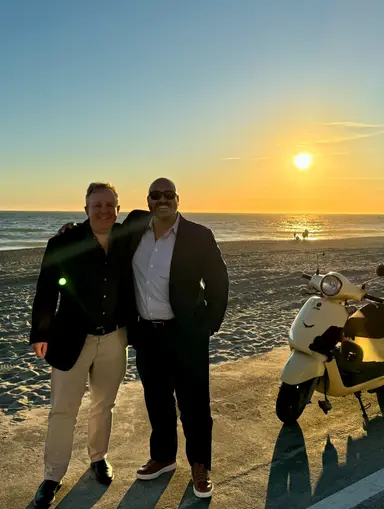 Two men in dark jackets standing on a beach at sunset next to a white scooter.