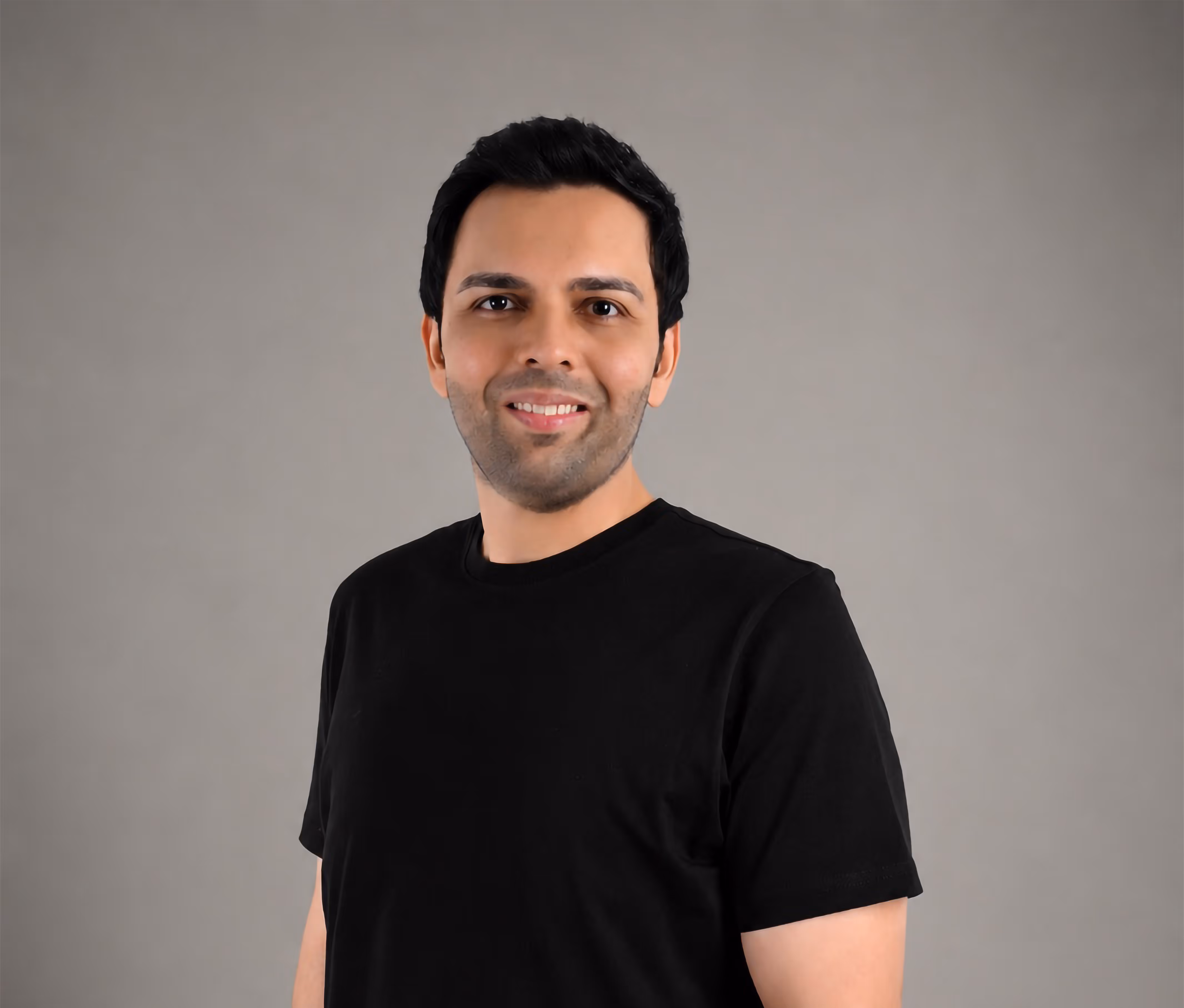 Smiling man with short black hair wearing a black t-shirt against a plain gray background.