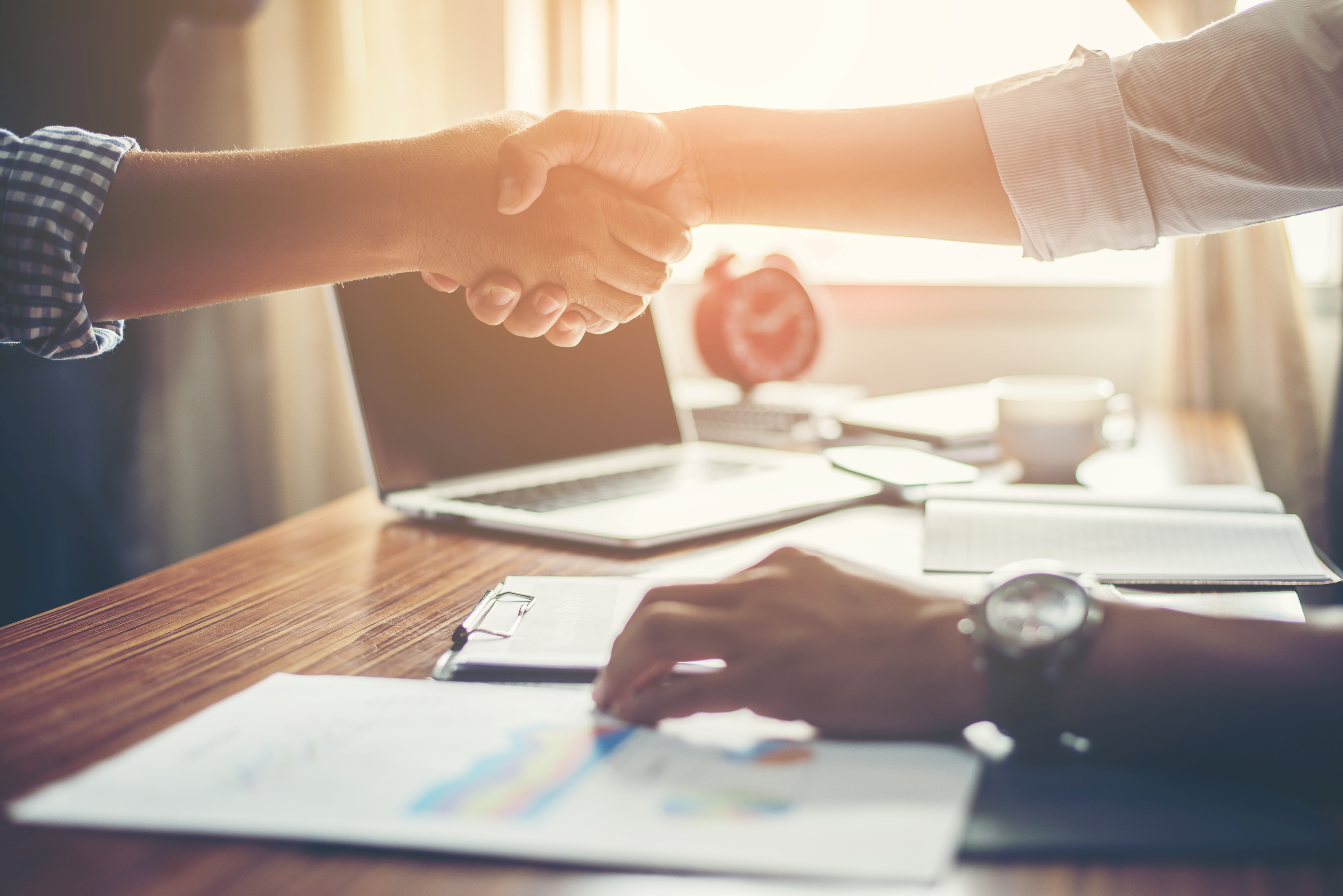 Two people shaking hands over a desk with a laptop, documents, and a coffee cup in sunlight.