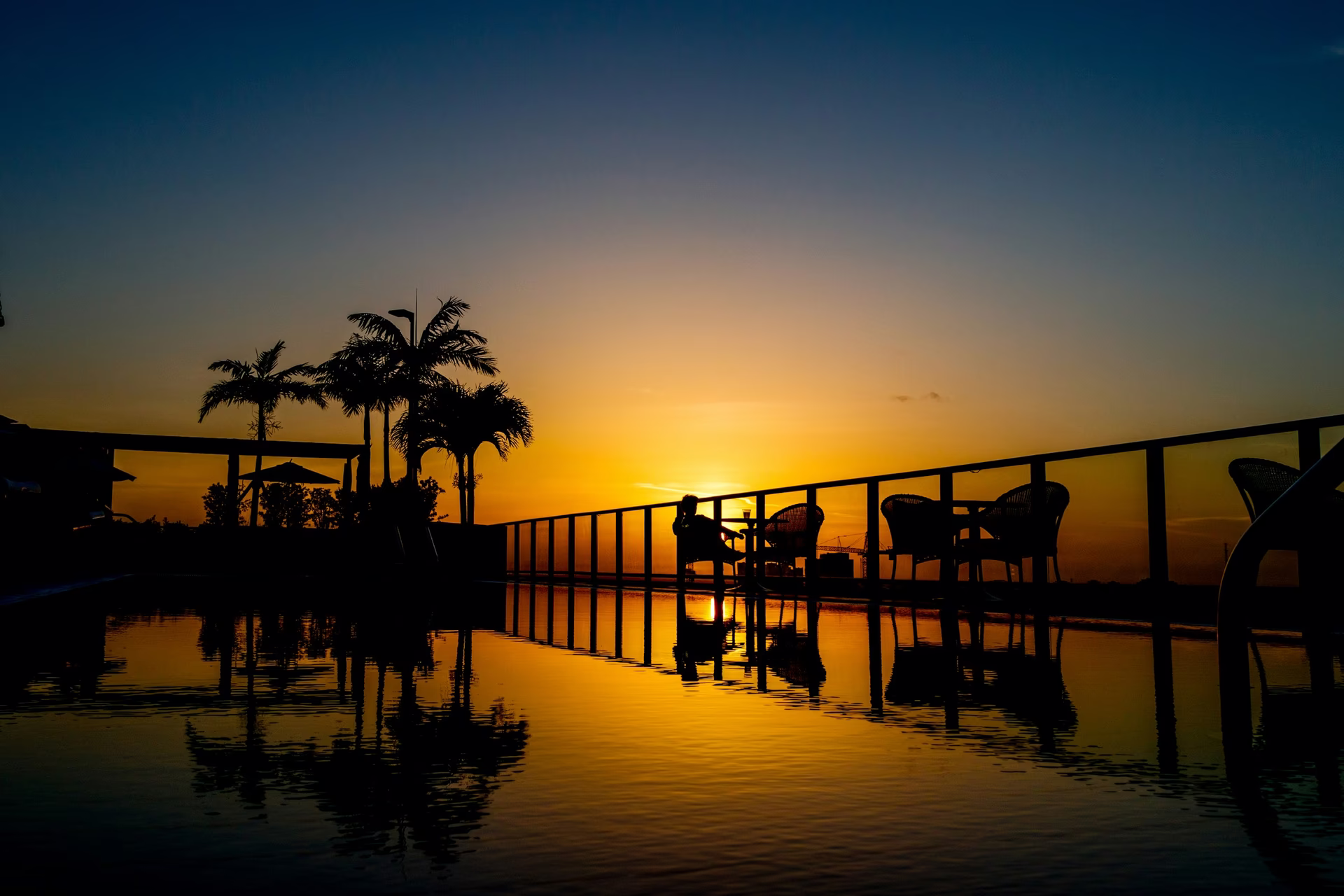 Silhouette of a person sitting by a pool with palm trees at sunset reflecting in water.
