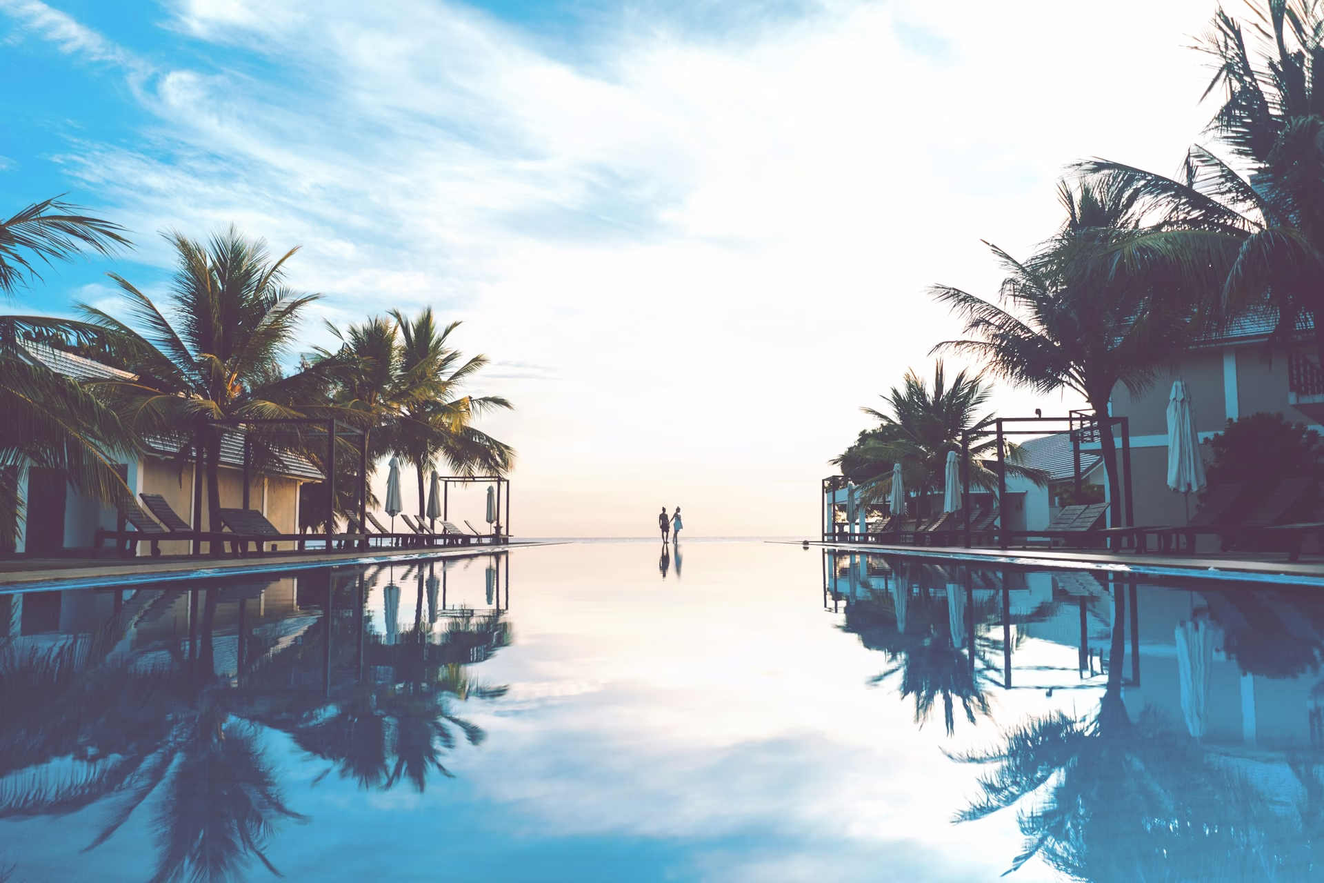 Couple standing at a luxury resort infinity pool surrounded by palm trees at dusk.