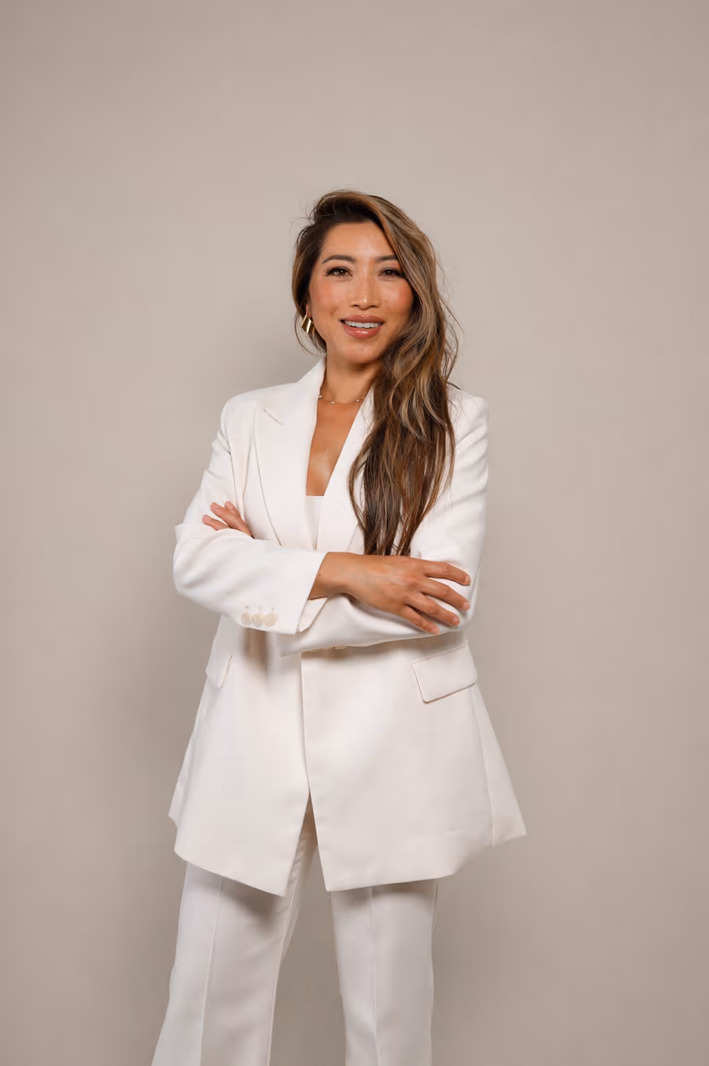 Dr.Nency with long hair wearing a white suit, smiling with arms crossed against a plain background.