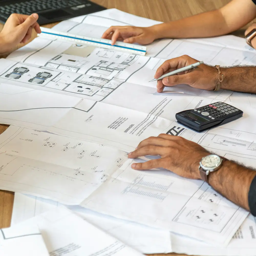 Personer arbejder på arkitekttegninger med lineal, pen og lommeregner på et bord.