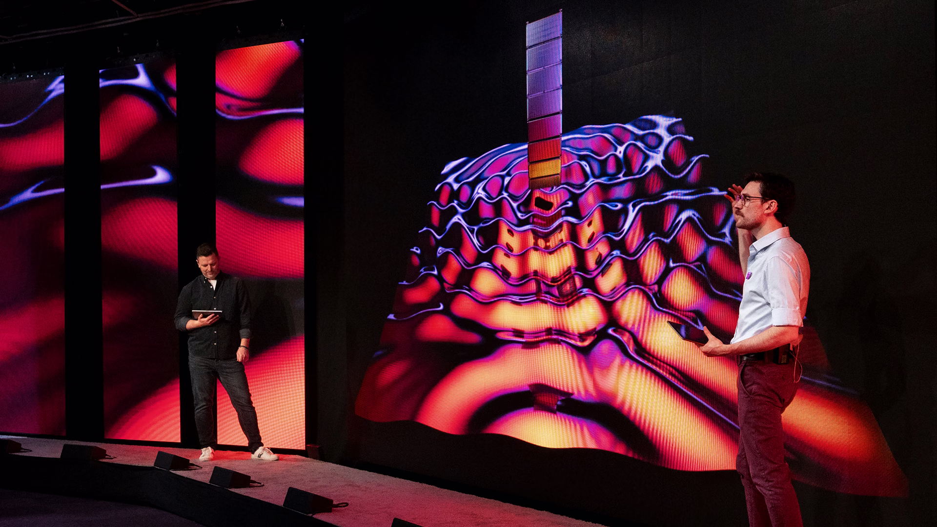 Presenter speaking beside a large colourful generative visual at the event.
