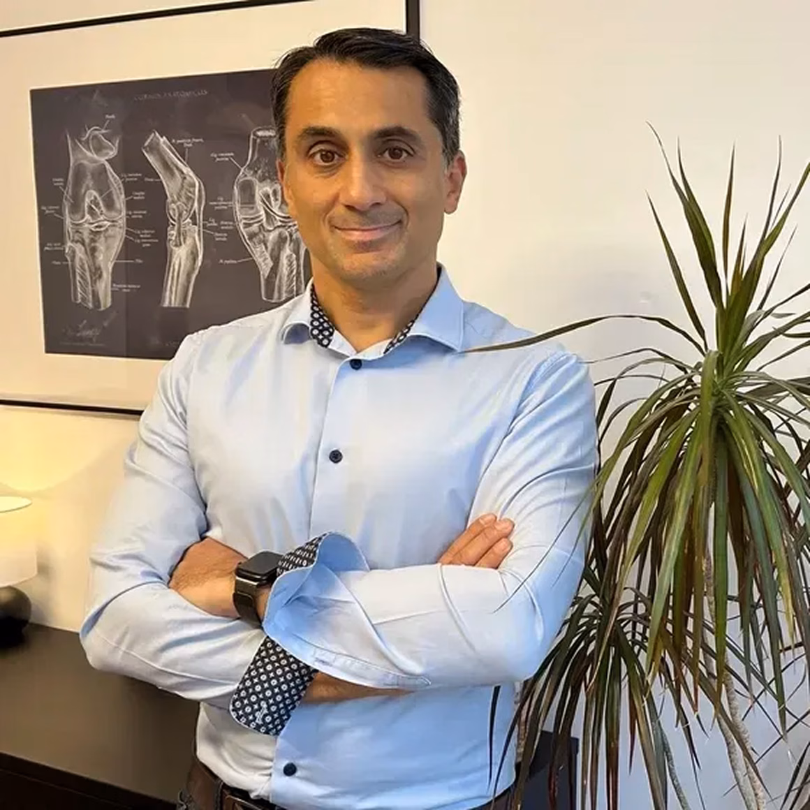 Man in light blue shirt with arms crossed standing next to a plant and anatomical knee joint poster on the wall.