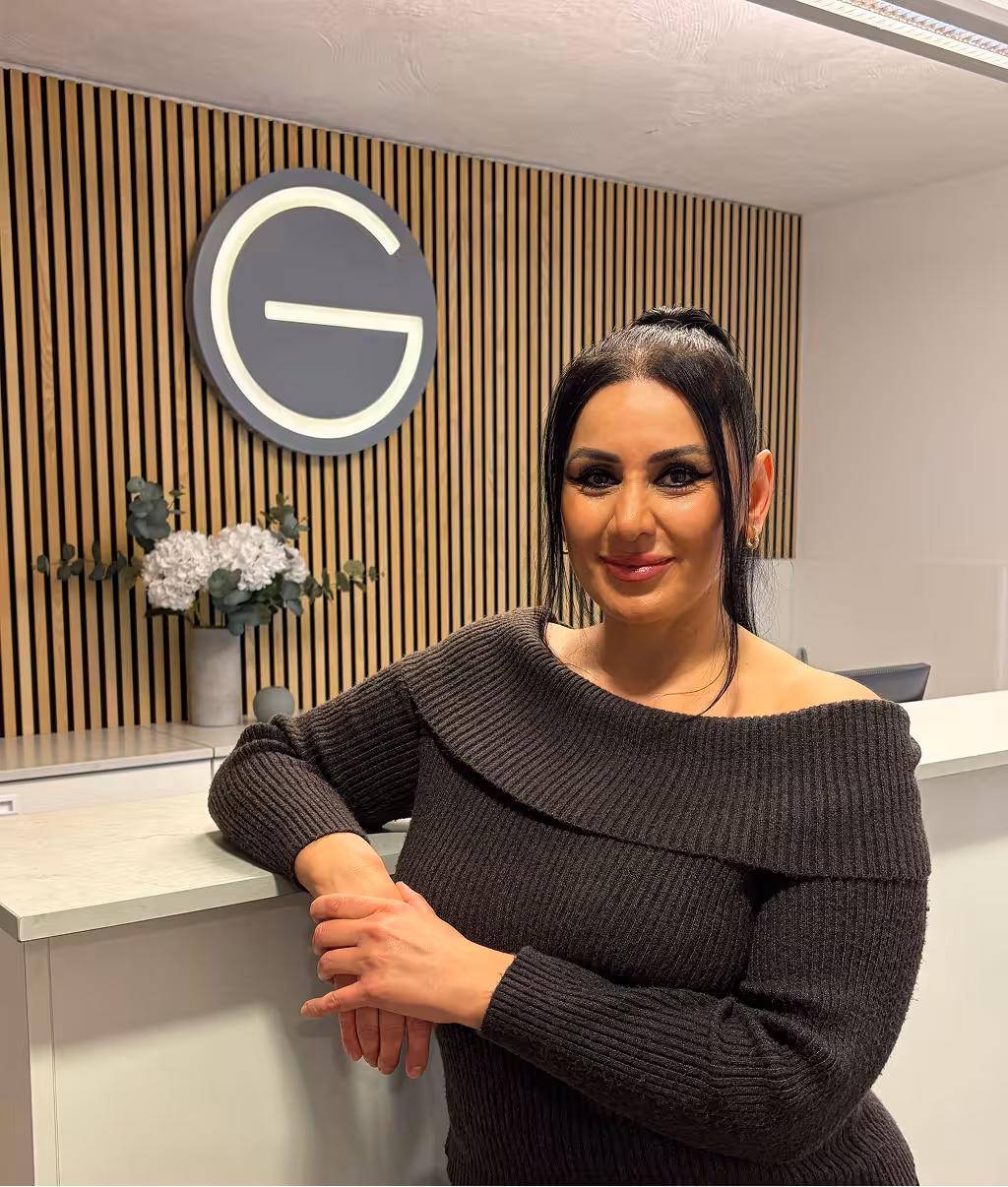 Woman with dark hair in a black off-the-shoulder sweater leaning on a white desk in a modern office with wooden slat wall and illuminated letter G logo.