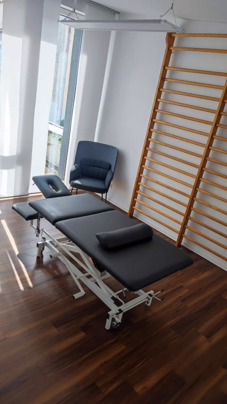 Treatment room with a black padded massage table, a gray cushioned chair, and wooden wall-mounted exercise bars.