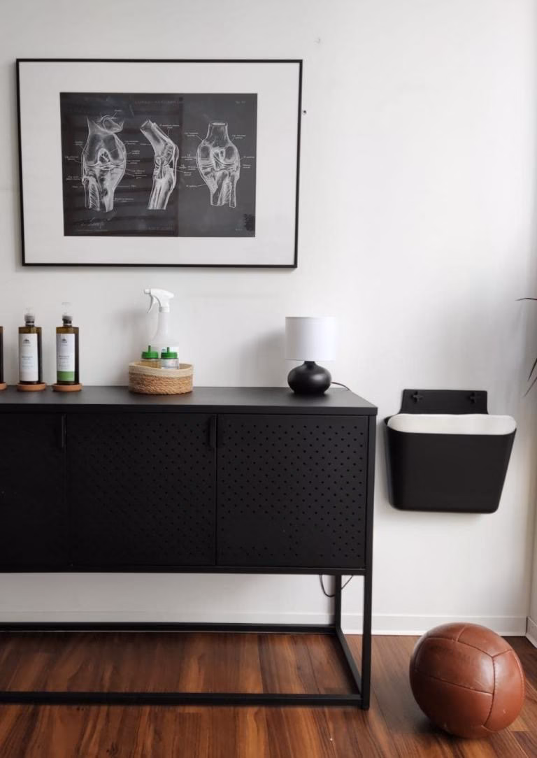 Black cabinet with skincare bottles, a small lamp, and a framed anatomical knee joint illustration on a white wall, next to a brown leather medicine ball on a wooden floor.