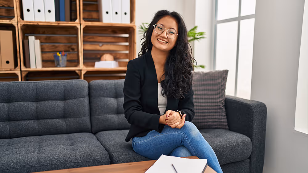 Professional woman sitting on gray sofa, wearing black blazer and glasses, smiling at camera