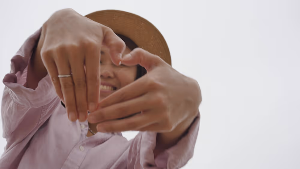 Woman in pink shirt making heart shape with hands while smiling outdoors