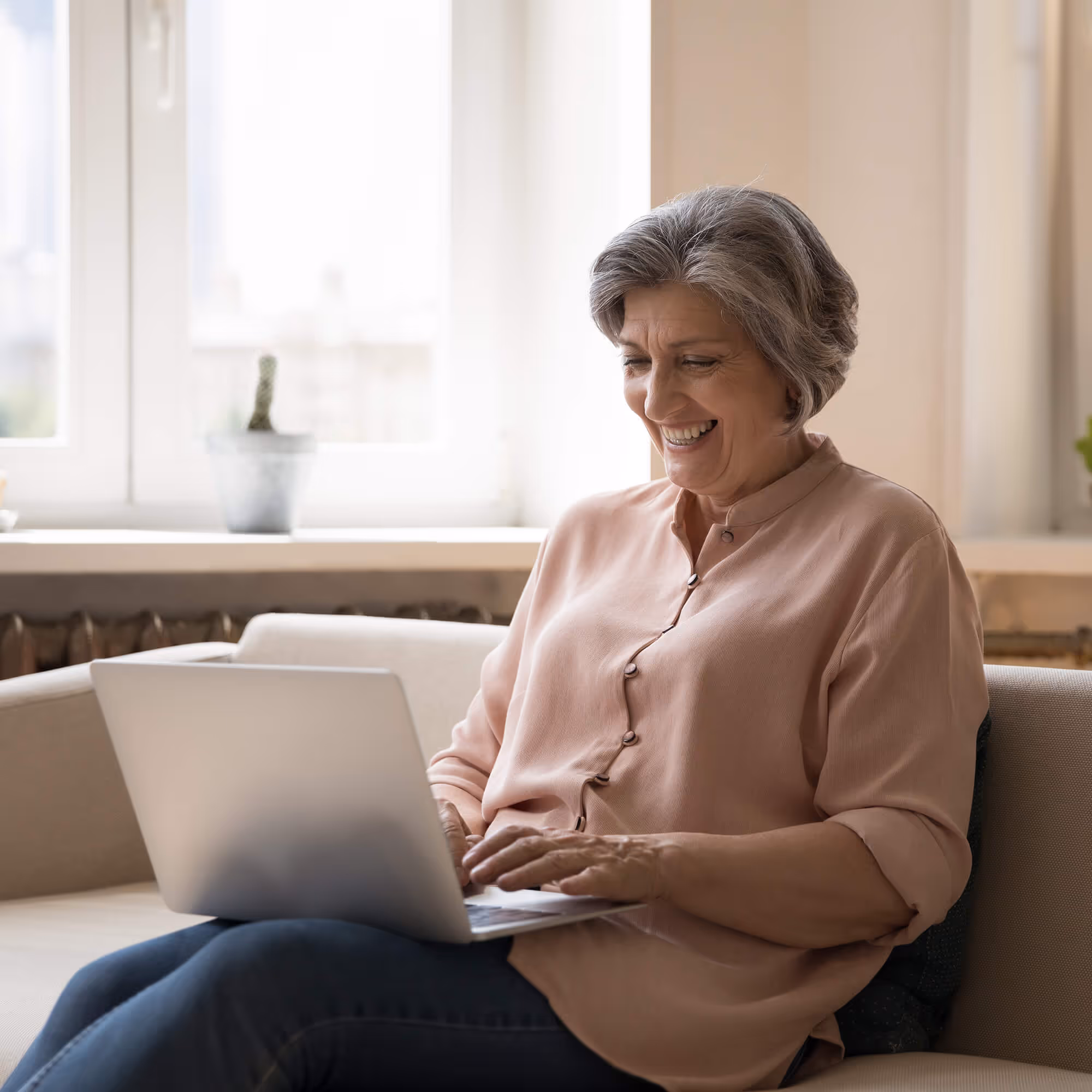 Mature woman smiling while using laptop on couch by bright window