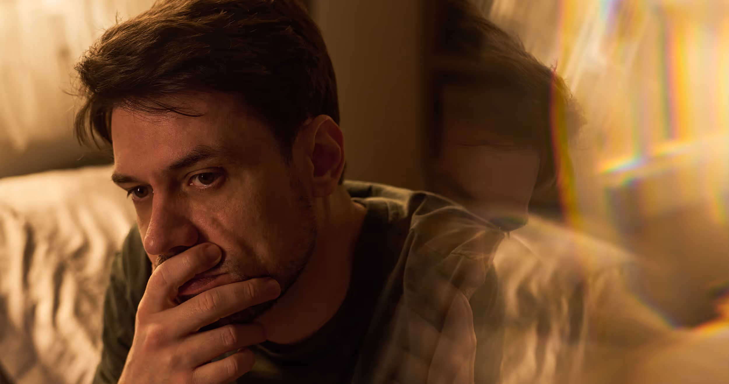 Man looks thoughtfully away with hand near mouth, warm interior lighting with rainbow prism effect
