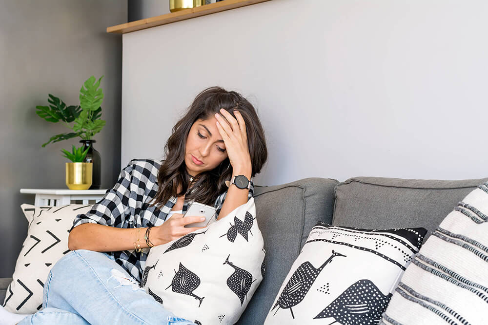 Woman sitting on couch looking at smartphone with concerned expression, head in hand.