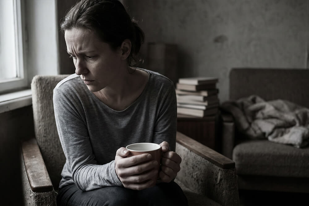 Woman sitting alone holding cup, looking down pensively in dim room