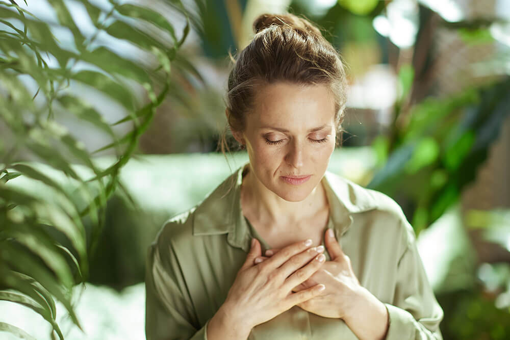 Woman in sage green shirt standing among green plants, looking down peacefully with hands on chest