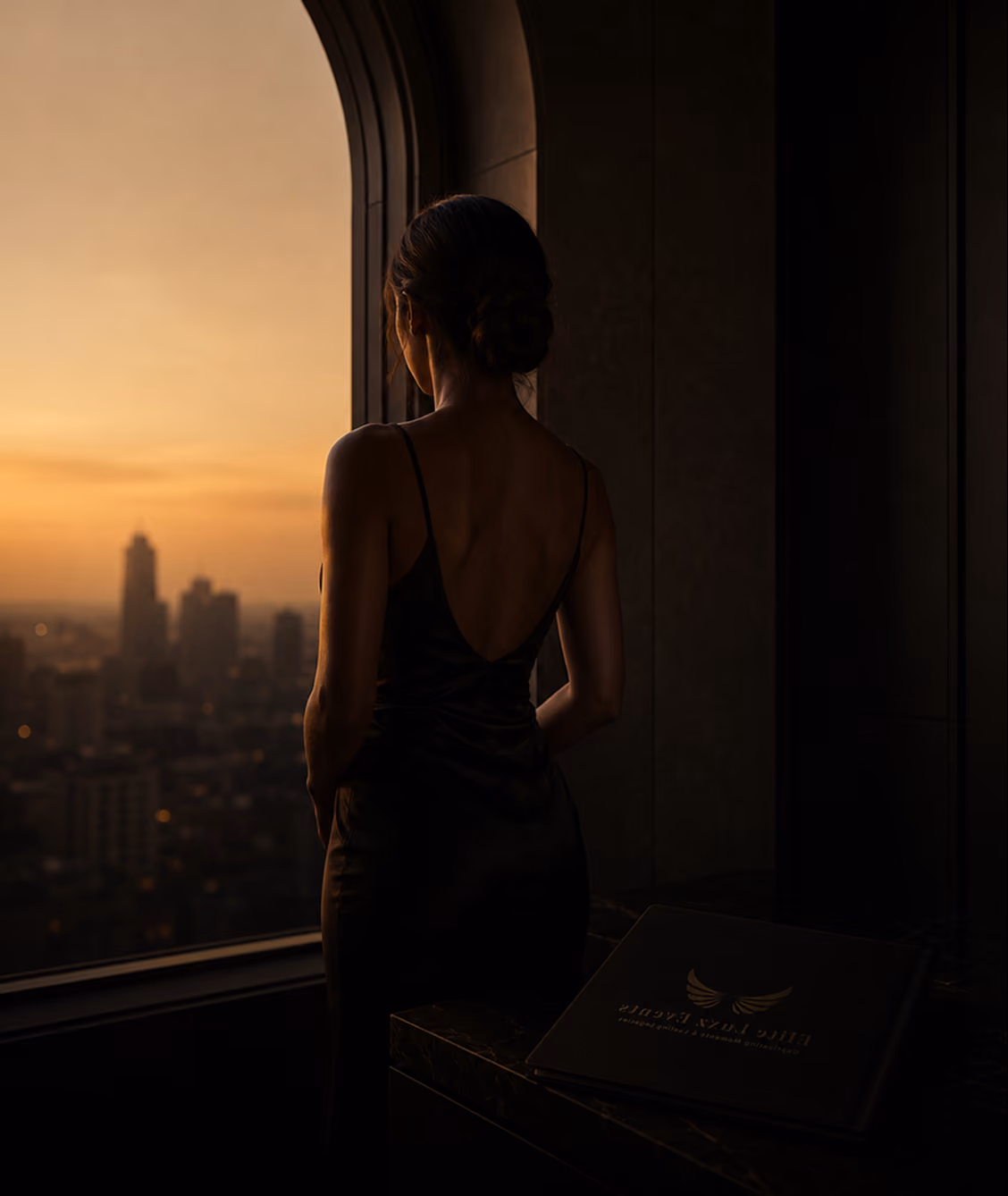Woman in an evening gown looking out over a city skyline at sunset, with an Elite Luxz Events branded book nearby