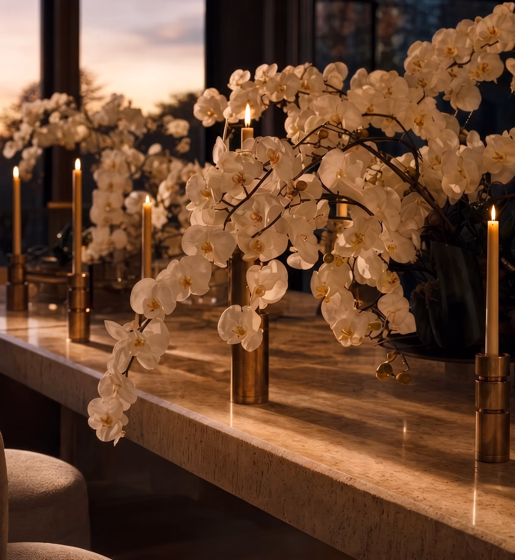 White orchids arranged on a marble table beside lit taper candles at sunset.