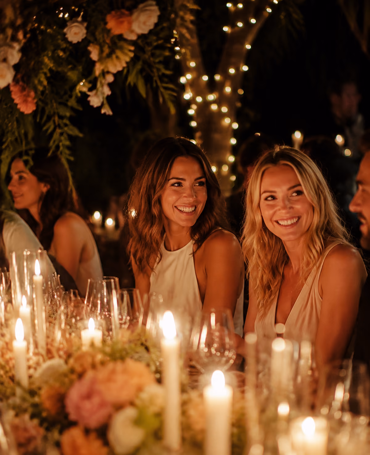 Two smiling women at a candlelit dinner table with flowers and fairy lights in the background.