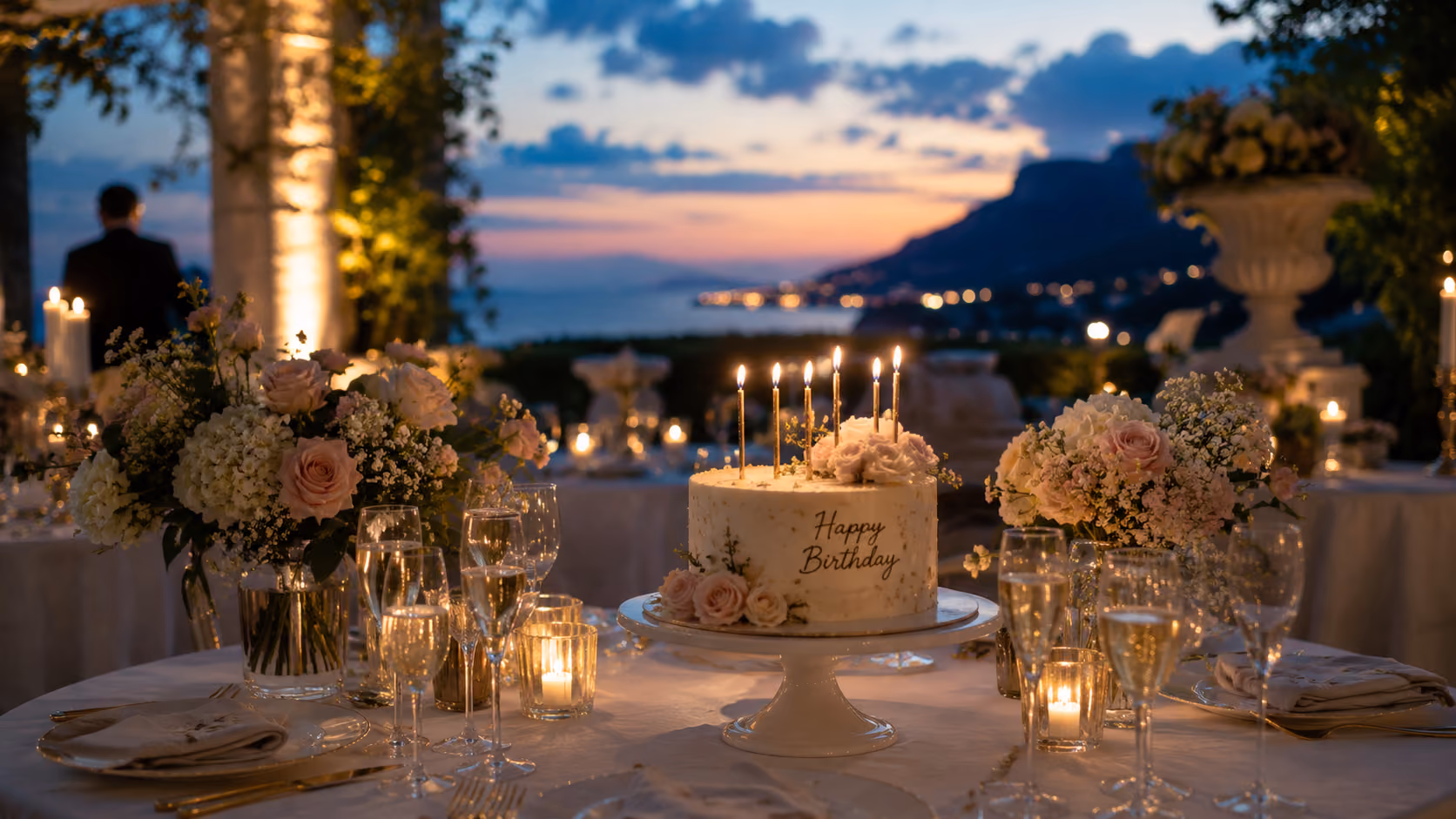 Birthday cake with candles on a table decorated with flowers and glasses at sunset outdoors.