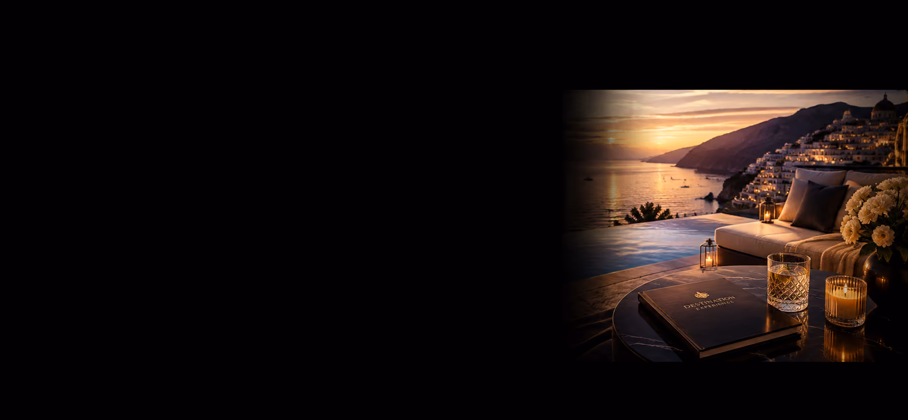 Sunset over a coastal town with lit buildings, overlooking a poolside table with drink, candle, and flowers.