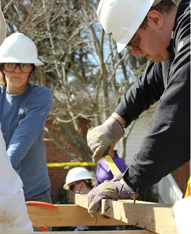Construction workers wearing helmets and gloves hammering nails into a wooden plank outdoors.