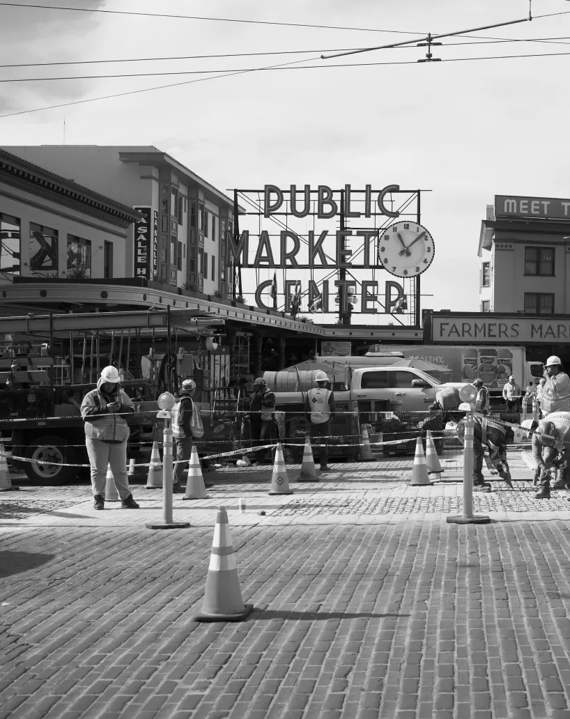 Construction workers on a cobblestone street in front of Public Market Center sign and Farmers Market entrance.