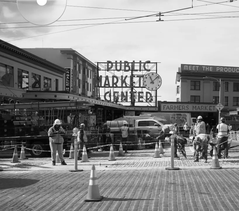 Construction workers on a cobblestone street in front of Public Market Center sign and Farmers Market entrance.