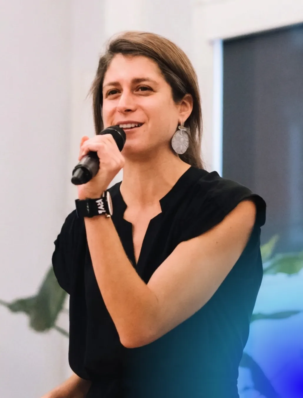 Woman with earrings speaking into a microphone, wearing a black top and a wristband.