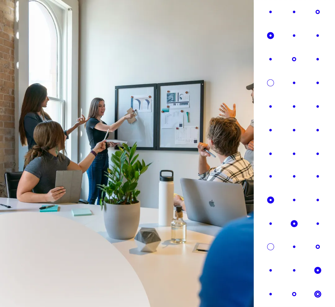 A group of coworkers discuss and point to papers on wall boards during a meeting in an office.