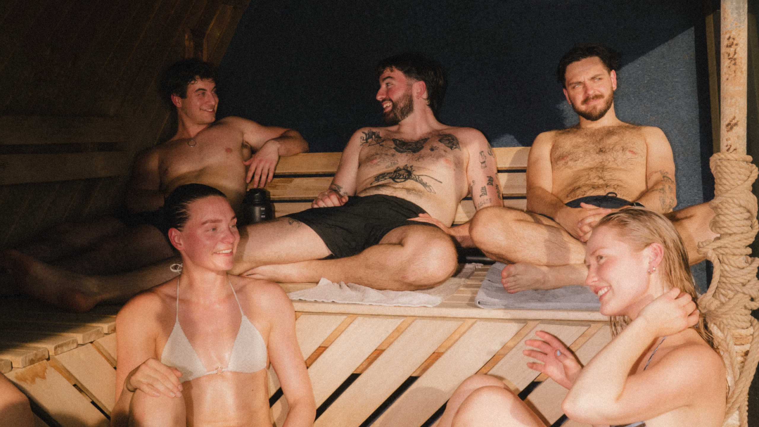 Five young adults relaxing and socializing in a wooden sauna, smiling and chatting together.