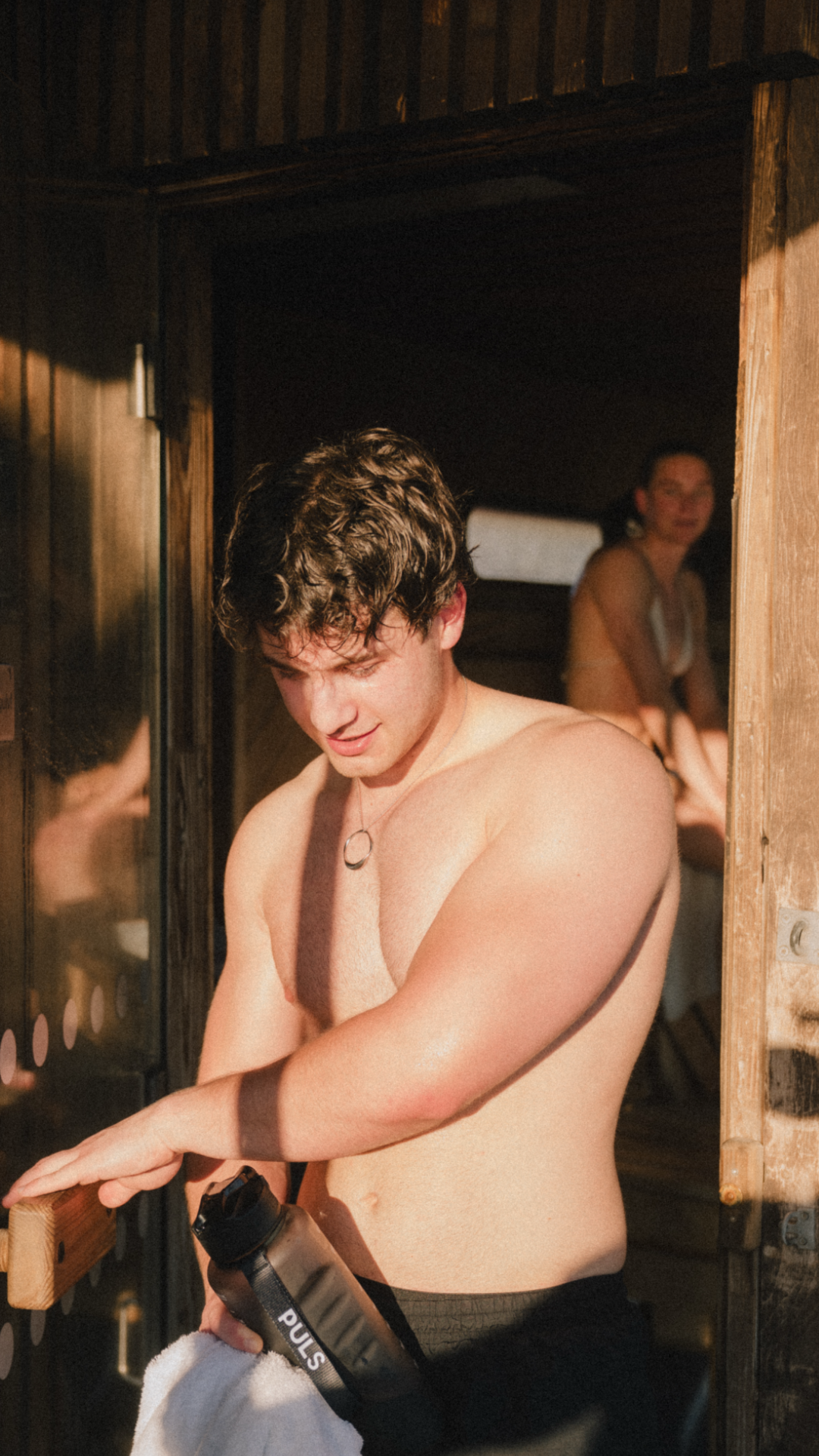 Shirtless man holding a water bottle and towel by an open wooden door with another person in the background.