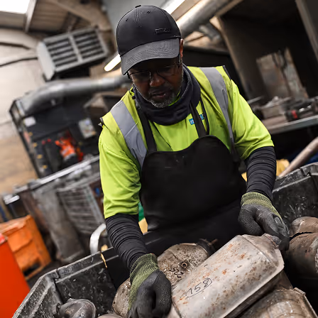 Worker in gloves and high-visibility shirt sorting scrap metal in industrial setting.