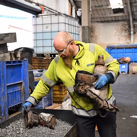 Man in a high-visibility jacket inspecting heavy metal machinery parts in an industrial warehouse.