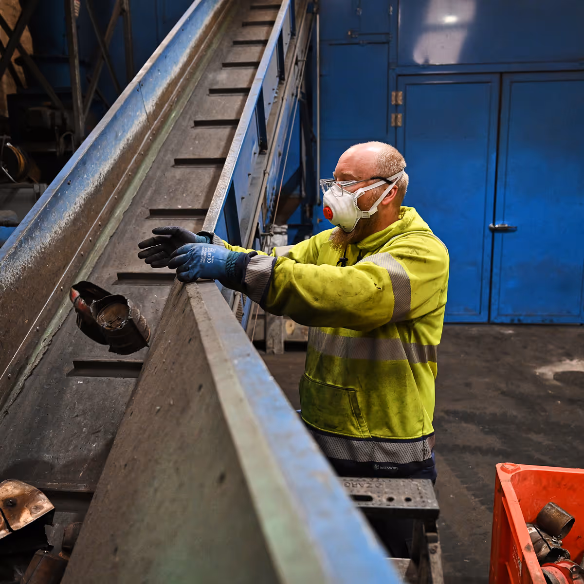 Worker in protective mask and gloves sorting metal pieces on a conveyor belt in an industrial facility.