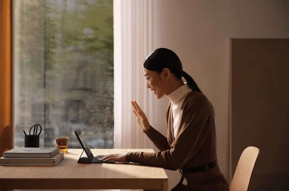 A woman smiling and waving on a video call from a sunlit wooden desk