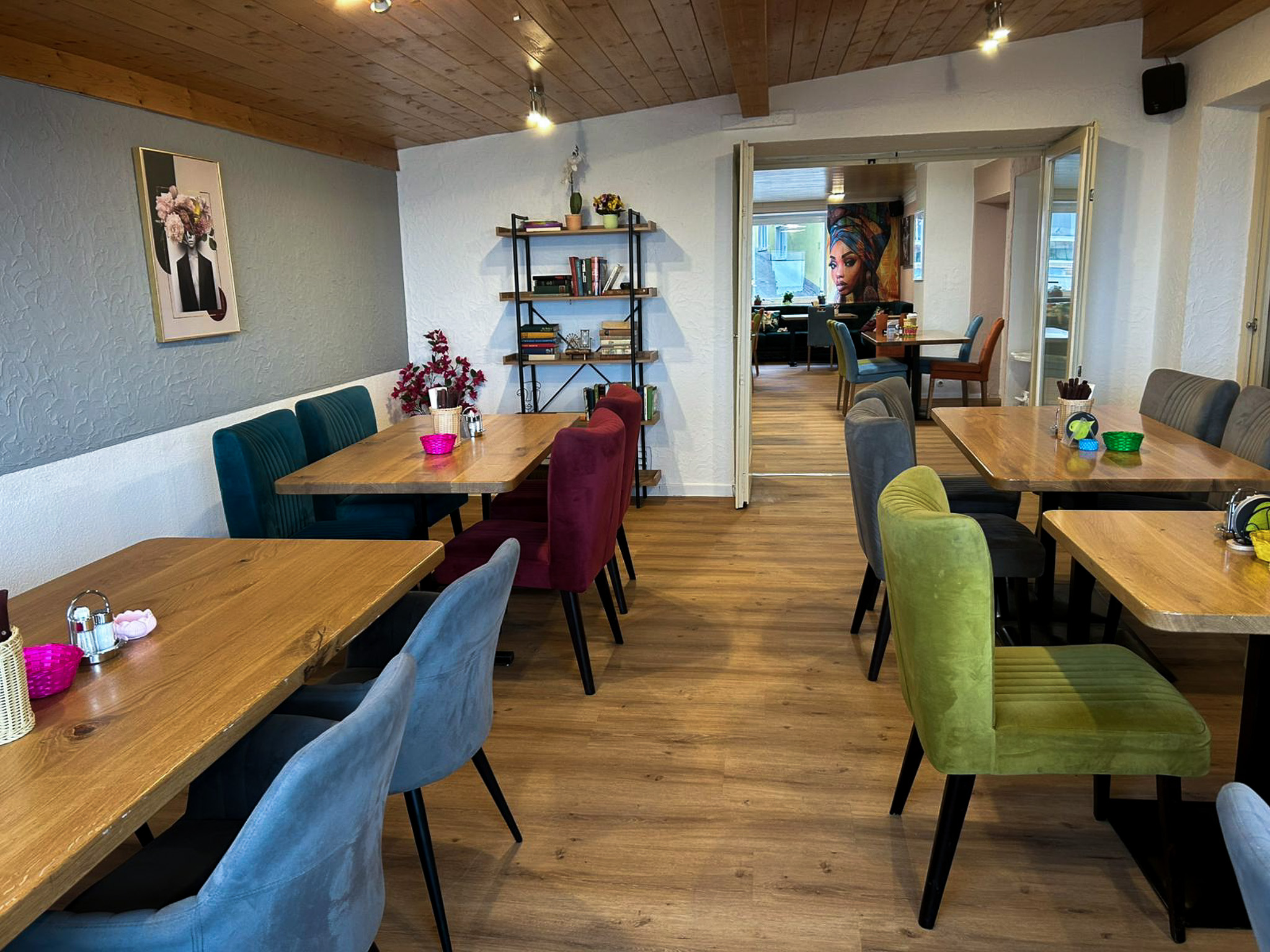 Gemütliches Restaurant mit bunten Stühlen, Holztischen, Bücherregal und Wandbild einer Frau.