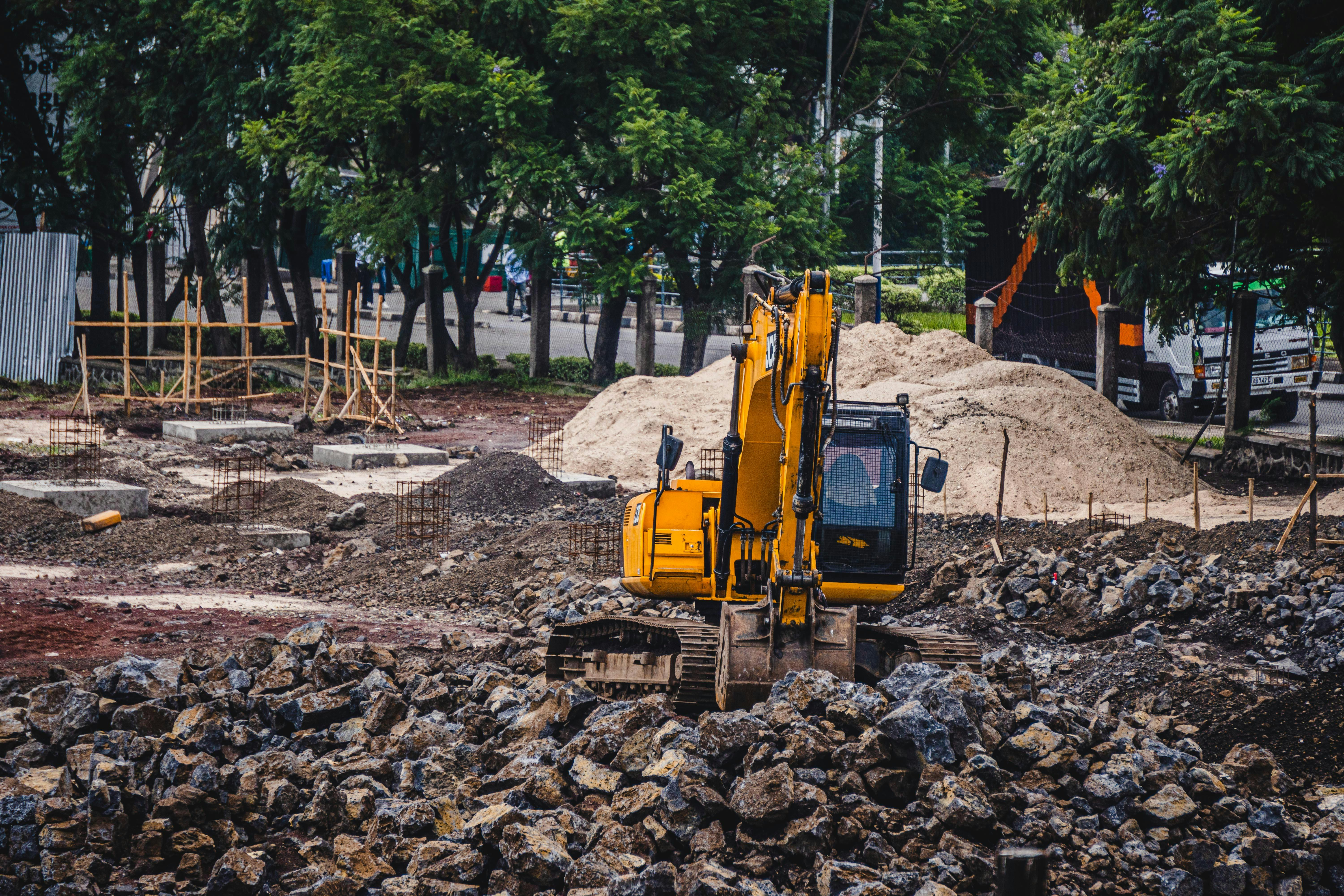 Yellow excavator working on a rocky construction site with piles of dirt and trees in the background.