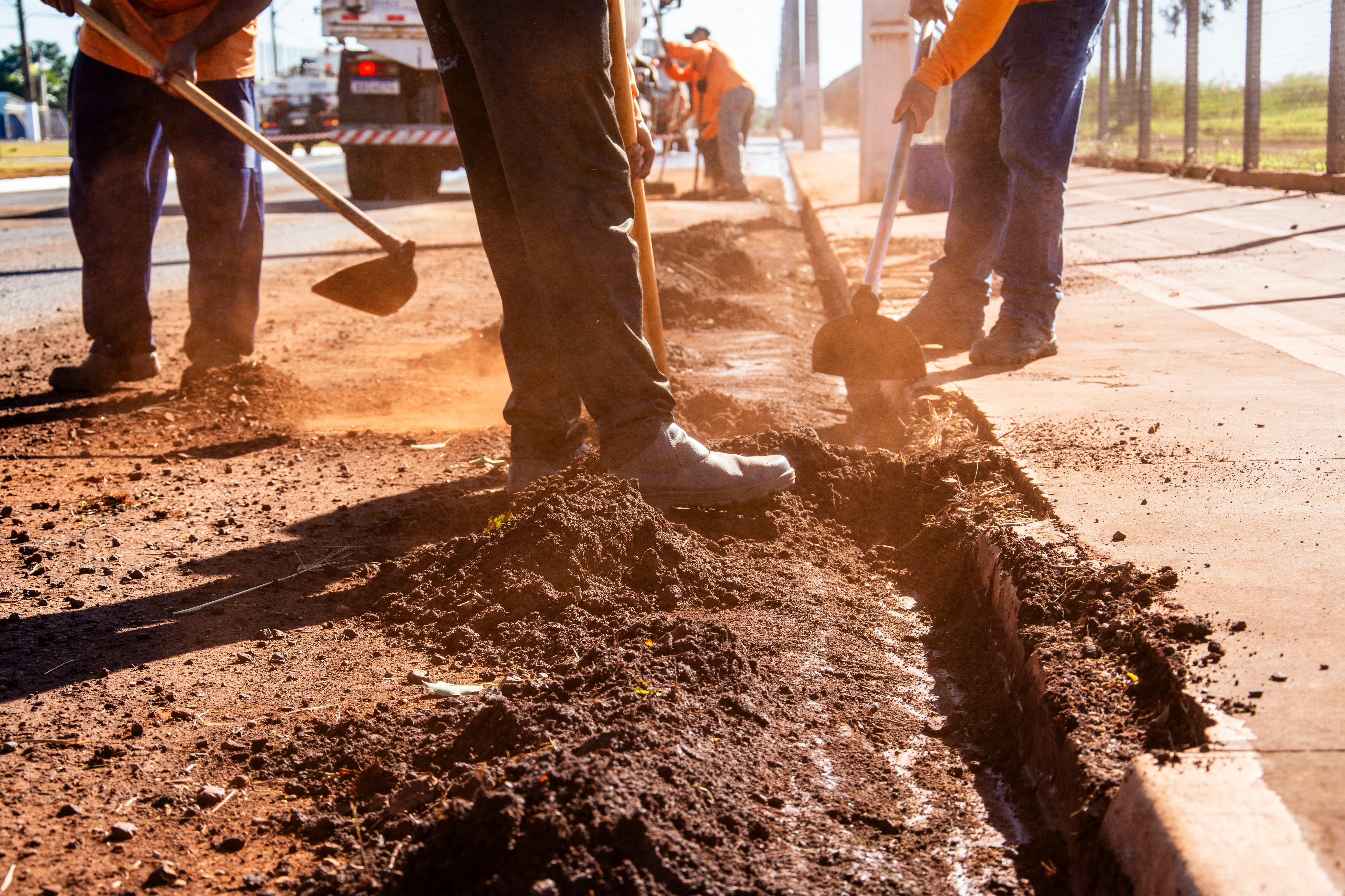 Workers digging and shoveling dirt along a road under bright sunlight.