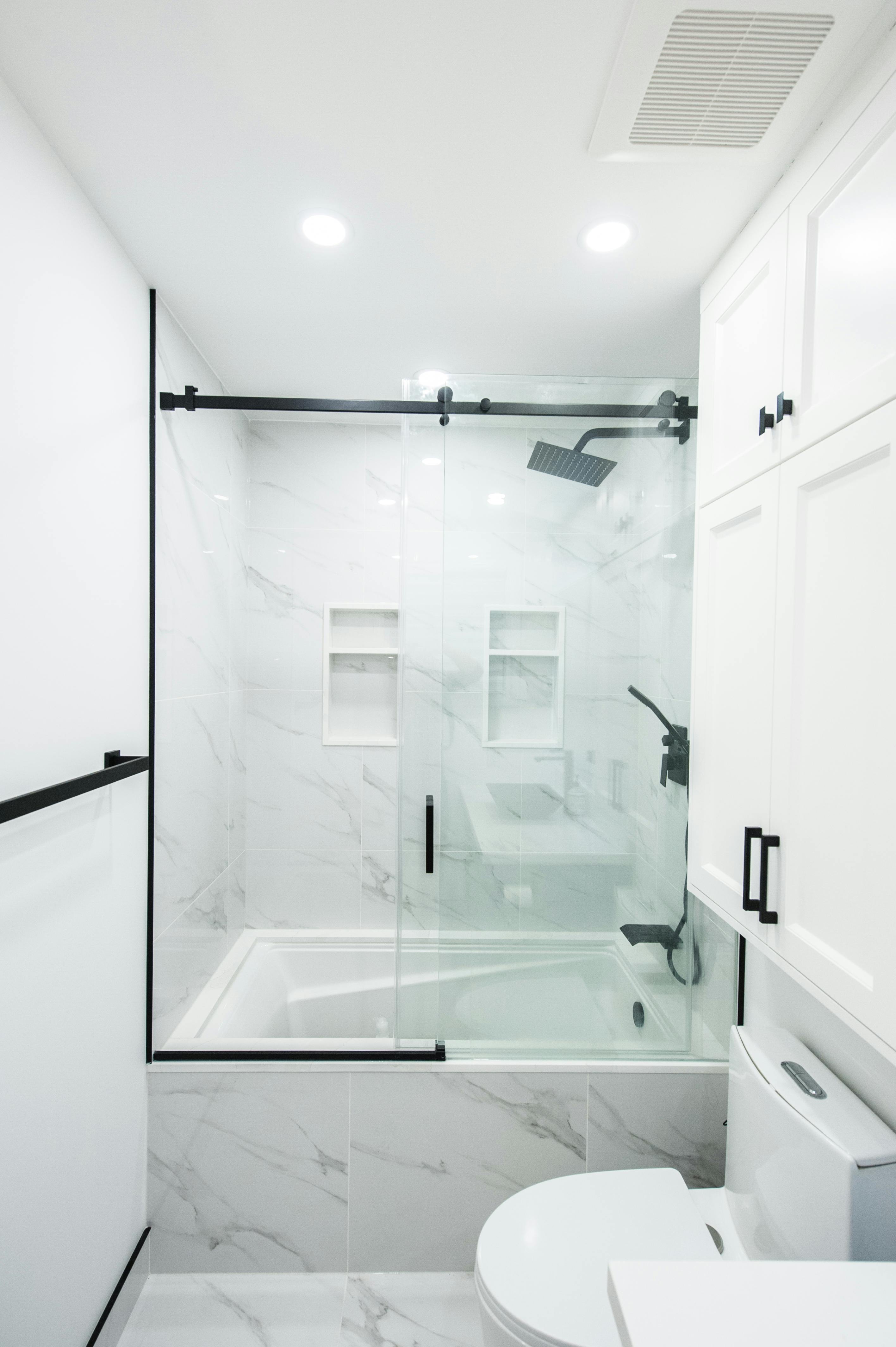 Modern white bathroom with marble tiles, glass shower enclosure with black hardware, and white cabinetry.